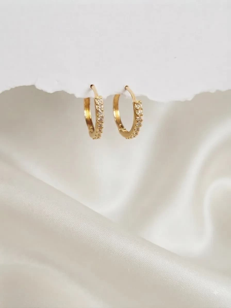 A pair of gold-plated huggie hoops lined with small clear stones, on ivory satin