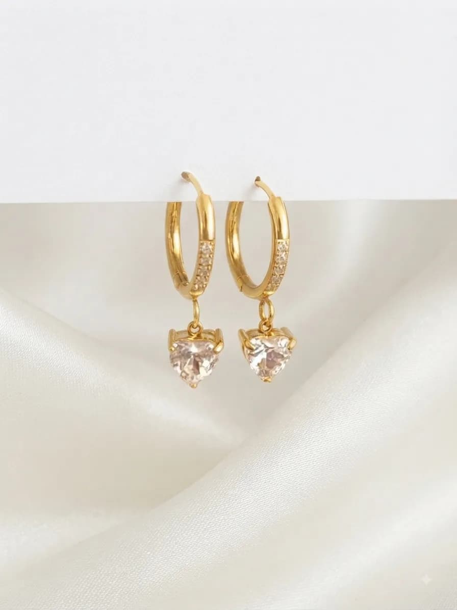 A pair of stone-set gold-plated huggies with a clear heart-shaped drop, on ivory satin