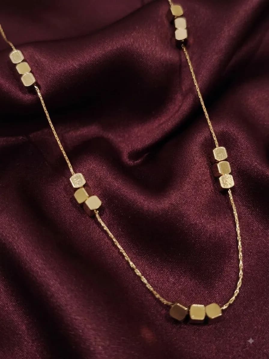 A fine gold-plated chain threaded with small square cube beads, laid on oxblood satin