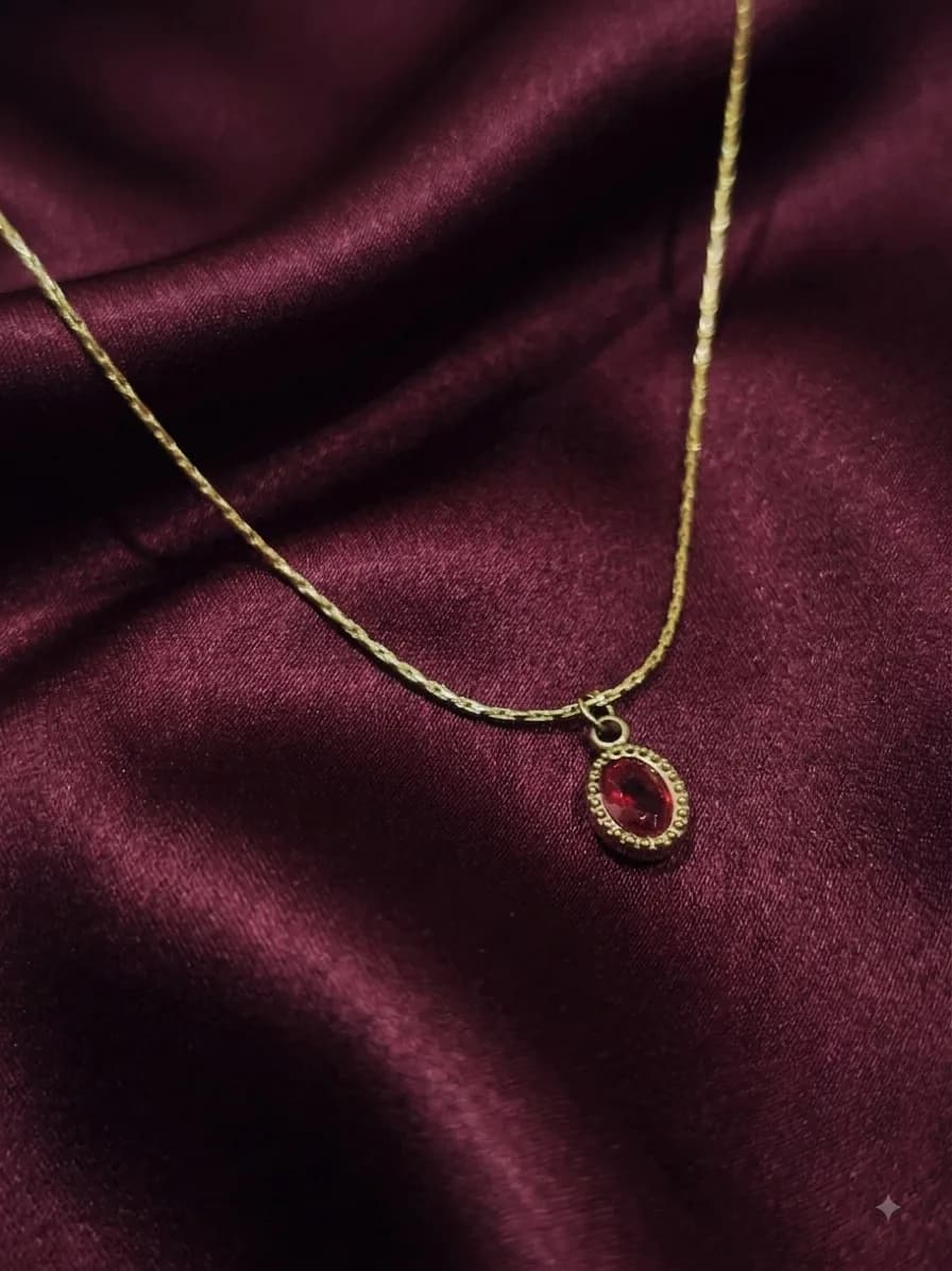 A gold-plated pendant holding a single oval ruby-red stone in a beaded bezel, on a fine chain against oxblood satin