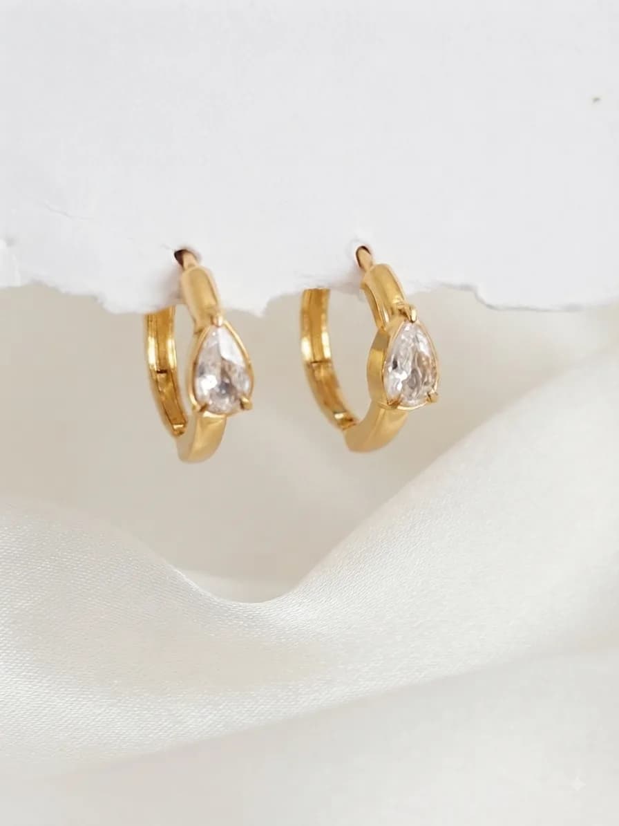 A pair of gold-plated huggies each holding a clear pear-cut stone, on ivory satin
